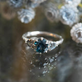 1.80ct Blue Montana Sapphire and Grey Spinel Three Stone Low Profile Evergreen Ring in Platinum on table with blue flowers